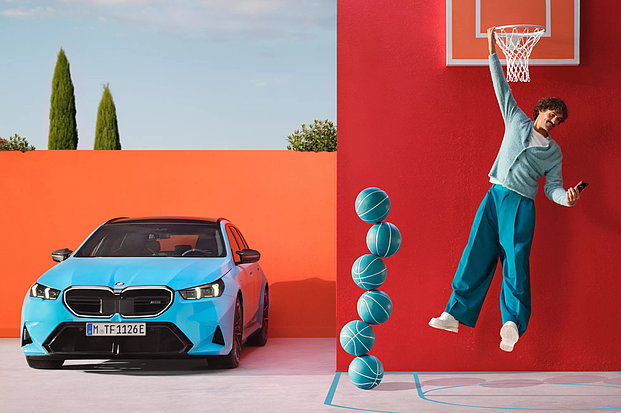 An einem Basketballkorb hängender Mann mit Handy in der Hand und BMW-Fahrzeug